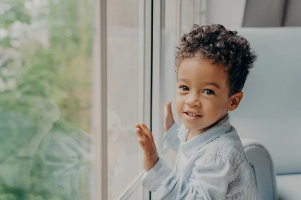 Lindo chico de pelo rizado de raza mixta en camisa de color azul claro junto a una ventana grande — Foto de Stock