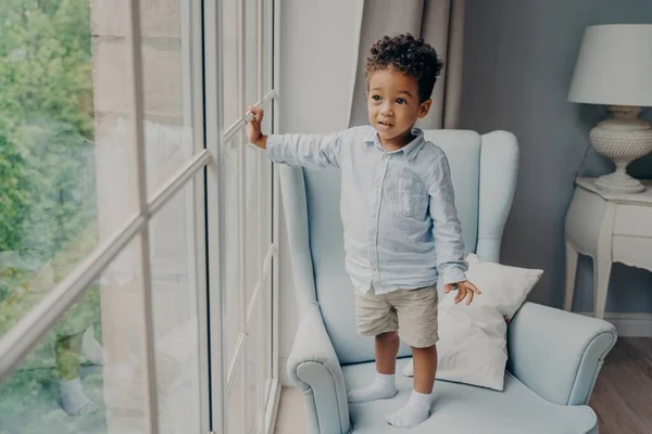 Curioso niño afroamericano mirando a través de la ventana mientras está de pie en el sillón en casa — Foto de Stock