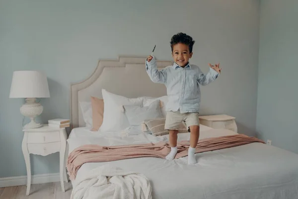 Alegre niño afroamericano pequeño saltando en la cama en casa y sonriendo — Foto de Stock