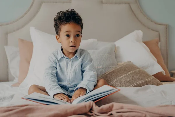 Lindo rizado afro americano pequeño niño sentado en cama con libro — Foto de Stock