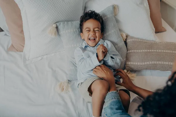 Adorable hijito acostado en almohadas en la cama y riendo mientras mamá le hace cosquillas — Foto de Stock