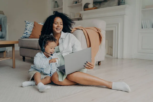 Niño pequeño feliz usando auriculares mientras habla en línea en el ordenador portátil, sentado en el suelo con mamá — Foto de Stock
