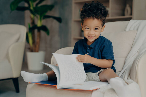 Cute little afro american boy sitting in armchair with story book
