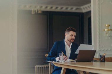 Akıllı takım elbiseli ciddi bir işadamı, ofis masasındaki kağıt dökümanlara yoğunlaşmış olarak not defterinde yazar. Bilgisayarlı cam bardak su dinleme bilgileri için kablosuz kulaklık kullanır..