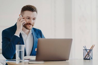 Genç işadamı ofis masasının arkasında dizüstü bilgisayarın önünde telefonla konuşuyor.