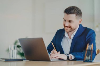 Resmi takım elbiseli şirket çalışanı masaüstünde oturup ofis içi dizüstü bilgisayara odaklanır. Kalem saatleri ile ilgili bilgileri yazar. Özel web sitesi araştırmaları yapar.