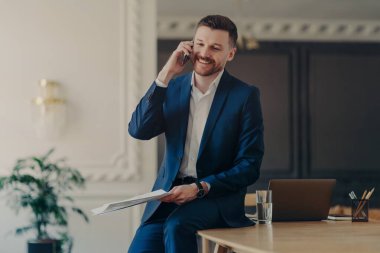 Resmi takım elbiseli girişimcinin kapalı çekimleri telefon görüşmesi yapıyor. Çalışma konularını partnerle tartışıyor. Masaüstüne yaslanıyor ve sunuma hazırlanıyor.