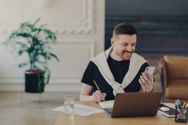 Mutlu sakallı genç adam kapalı alanda modern aletlerle çalışır internet sitesinden notlar yazar not defterine notlar yazar birlikte çalışan rahat bir yerde akıllı telefon pozları verir. Hippi öğrenci.