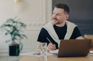 Neşeli erkek öğrenci ev ödevi için dizüstü bilgisayarla bilgi arıyor. Makale yazıyor. Ortak çalışma alanında oturuyor. Mutlu bir gülümsemeyle bakıyor.