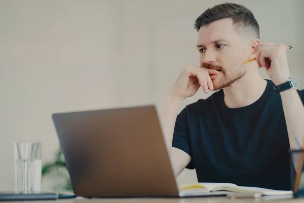 Grave studente uomo barbuto gode di studiare processo ha espressione premurosa lavora freelance vestito con casual t shirt nera annota utilizza moderni computer portatili posa in spazio di coworking — Foto Stock