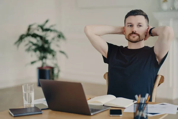 Il collaboratore esterno maschile pensieroso prende la pausa tiene mani dietro pose di testa a lavori fatti col personal computer su computer portatile ha l'espressione premurosa pensa a piani futuri la settimana vestita di t-camicia nera. — Foto Stock