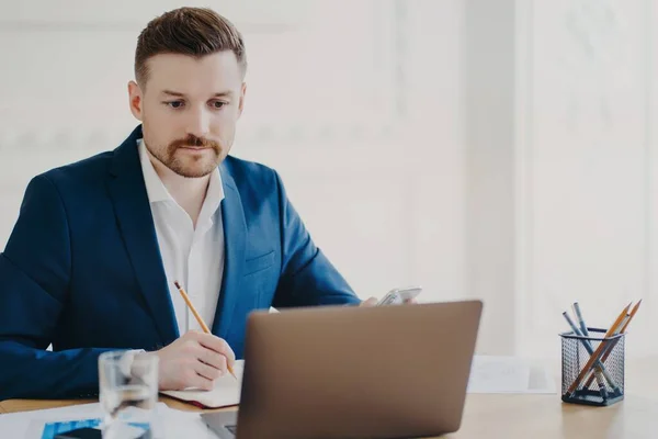 El vendedor barbudo serio implicado en el proceso de trabajo toma notas con lápiz mira atentamente a la computadora portátil lleva traje formal azul posa en el espacio de coworking escribe plan de organización — Foto de Stock