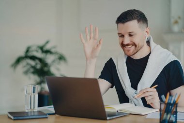 Neşeli sakallı adamın video konferansı var. Bilgisayarda avuç içi bilgisayar var. İş yerinde kablosuz kulaklık kullanıyor.