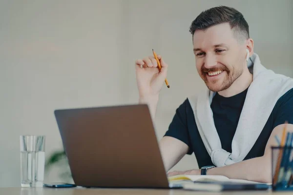 El hombre barbudo feliz prepara el trabajo de curso de papel funciona de forma remota desde casa relojes webinar utiliza conexión a Internet de alta velocidad portátil moderno y auriculares conversaciones con el socio en línea. Tiempo de trabajo. — Foto de Stock