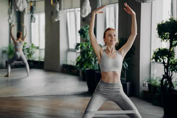 Atletica sottile rossa istruttore di fitness si siede facendo squat con pilates elastico elastico di resistenza sulle gambe — Foto Stock