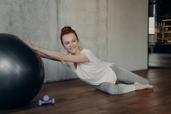 Sorridente donna zenzero sportivo in abbigliamento sportivo posa sul pavimento in legno con palla fitness argento — Foto Stock