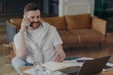 Neşeli serbest çalışan bir adam telefonla bilgi yazar not defterine not alır dizüstü bilgisayardaki iç dizüstü bilgisayar pozlarına dikkatle bakar. Teknoloji kullanıcısı