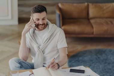 Süveterle kaplı beyaz tişörtlü gülümseyen adamın portresi sandalyede oturuyor ve telefonda konuşuyor.