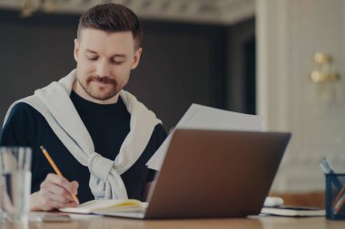 Günlük giysiler içinde serbest çalışan bir erkek ofis masasında otururken belgelerle çalışıyor.