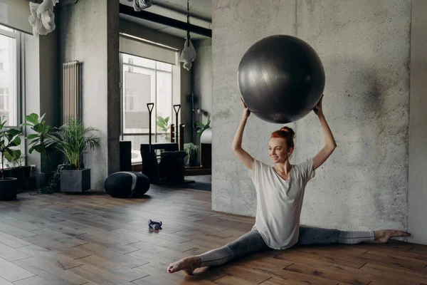 Bella donna fitness in posizione divisa tenendo palla esercizio sopra la testa e godendo di allenamento pilates — Foto Stock