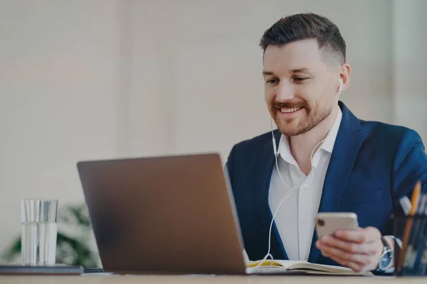 Trabalhador de escritório masculino feliz usa tecnologias modernas poses na área de trabalho detém telefone móvel faz chamada de vídeo via laptop usa fones de ouvido vestidos formalmente tem expressão feliz relógios webinar de negócios — Fotografia de Stock