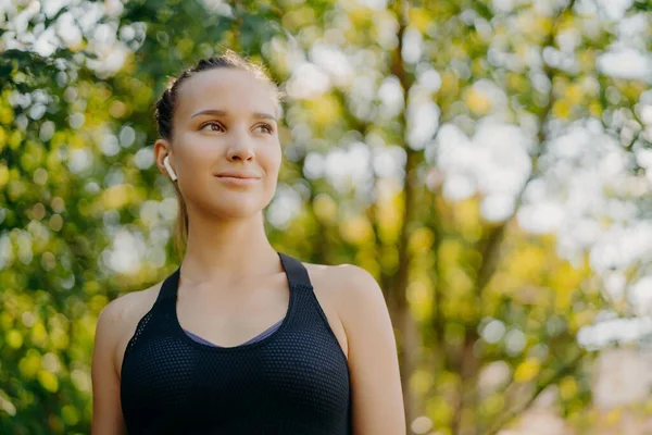 Aangename bedachtzame donkerharige vrouw in sportkleding kijkt weg met blije uitdrukking staat in groen park na joggen luistert favoriete muziek via draadloze oortelefoons — Stockfoto