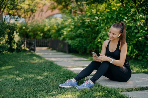 Mulher atlética agradável em roupas esportivas sorri alegremente surfa telefone celular enquanto ouve música através de fones de ouvido sem fio faz pausa após o treino matinal ensolarado usa aplicativo para rastreamento de atividades — Fotografia de Stock