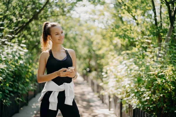 Plan horizontal de la femme sportive active utilise smartphone pour vérifier les résultats après le jogging habillé en tenue active bénéficie d'une journée chaude et ensoleillée écoute de la musique via des écouteurs. Concept de mode de vie sain. — Photo