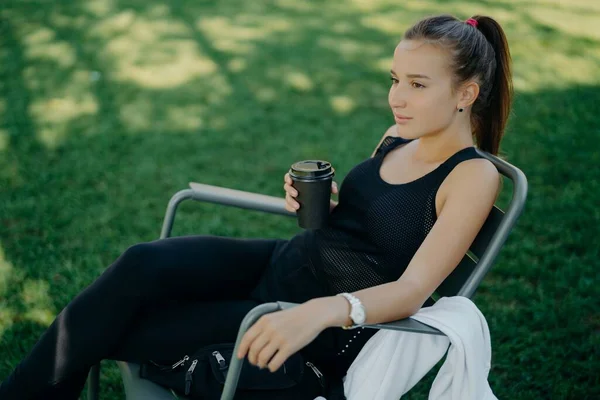 Outdoor shot of thoughtful dark haired beautiful female dressed in sports clothes drinks takeaway coffee spends free time in open air poses on chair at garden enjoys good weather breathes fresh air — Stock Photo, Image