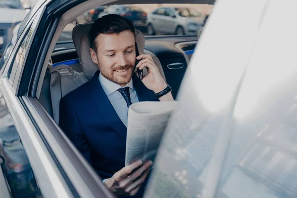 Good looking young executive manager in formal wear reads newspaper on backseat of car