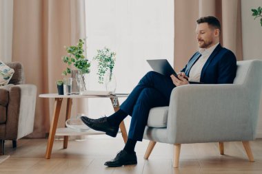 Resmi giyimde başarılı bir işadamı rahat koltuğunda oturur modern dizüstü bilgisayarlar kullanır çevrimiçi proje pozları ev içi analizler finansal göstergeler şirket gelişimini kontrol eder