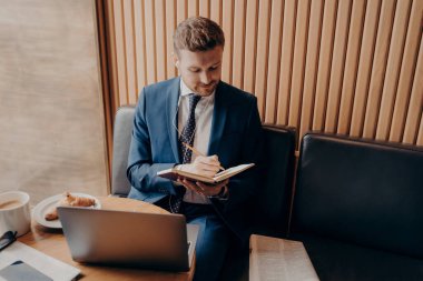 Genç sakallı işadamı kafede oturuyor ve internetteki internet ağları sırasında notlar yazıyor.