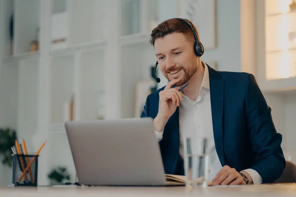 El operador masculino profesional trabaja en callcenter utiliza auriculares y conversaciones de computadora portátil en videollamada o evento virtual por webcam usa ropa formal posa contra el interior acogedor tiene reunión en línea — Foto de Stock