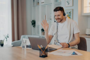 Kulaklıklı mutlu genç adam evden çalışıyor ve internet üzerinden iletişim kuruyor.