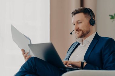 Düşünceli iş adamı elinde kağıtlar tutarken evde online projeyle çalışıyor.