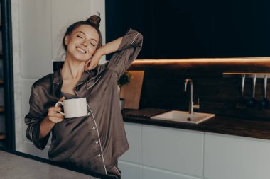 Güzel, rahat, genç bir bayan model. Mutfakta bir fincan kahveyle rahat pijamalar içinde.