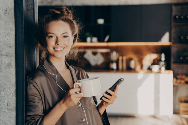 Lovely happy young woman having her morning coffee while checking new emails and notifications on smartphone