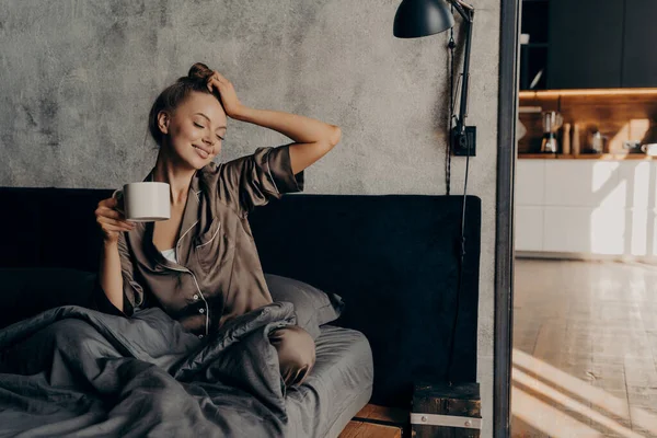Retrato de una joven mujer relajada en un elegante traje de casa despertando con una taza de café en la cama — Foto de Stock