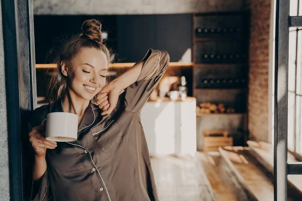 Feliz hermosa morena hembra estratando con taza de café en la mano mientras está de pie en la cocina — Foto de Stock