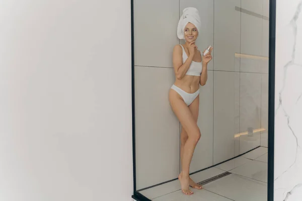 Όμορφη νεαρή γυναίκα που χρησιμοποιεί καλλυντικό προϊόν για υγιή επιδερμίδα και χαμογελά ενώ ποζάρει στο μπάνιο στο σπίτι — Φωτογραφία Αρχείου