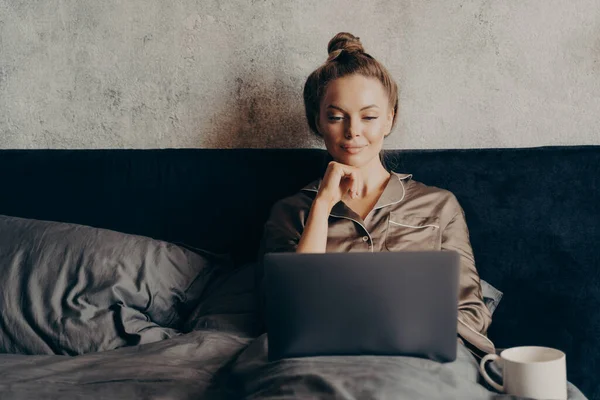 Positivo relaxado jovem mulher no pijama deitado na cama e usando laptop — Fotografia de Stock