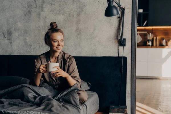 Alegre feliz sorrindo caucasiano fêmea em cetim pijama acolhedor segurando xícara de café — Fotografia de Stock