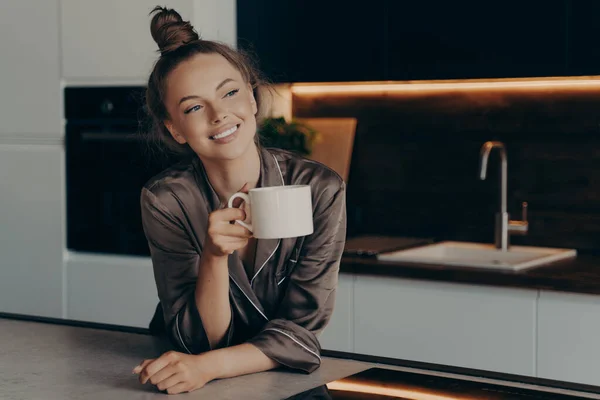 Felice sorridente giovane donna in pigiama di raso marrone godendo di caffè aromatico mattina in cucina — Foto Stock