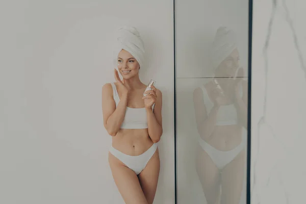 Όμορφη γυναίκα στέκεται στο μπάνιο μετά το πρωινό ντους και την εφαρμογή ενυδατική κρέμα στο πρόσωπο — Φωτογραφία Αρχείου