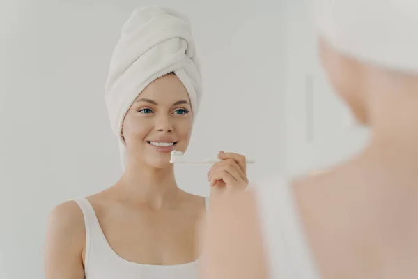 Healthy beautiful woman with towel on head after shower having toothy smile while gently brushing her teeth