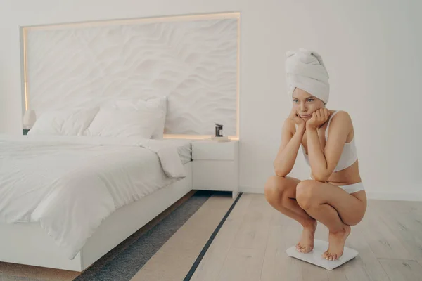 Young sad beautiful woman in white underwear standing on electronic smart scales in bedroom — Stock Photo, Image