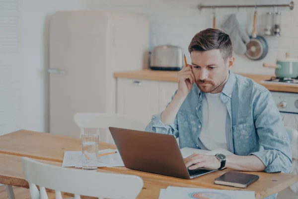 Homme d'affaires barbu concentré sérieux en vêtements décontractés travaillant sur ordinateur portable tout en étant assis à la table de cuisine — Photo