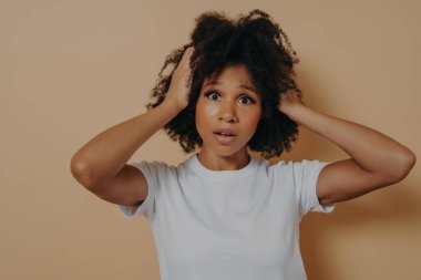 Endişeli, ağzı açık, başı bej arka planda kapalı, gergin bir Afrikalı kadın.