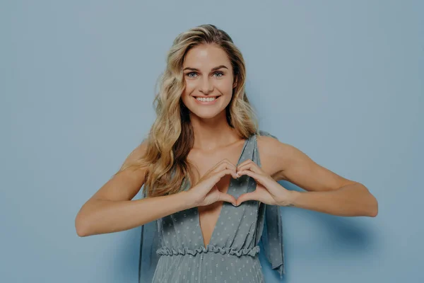 Happy lovely female with blonde long hair showing love symbol with her hands cupped in heart shape