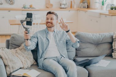 Kanepede otururken videoda barış işareti gösteren olumlu genç adam.